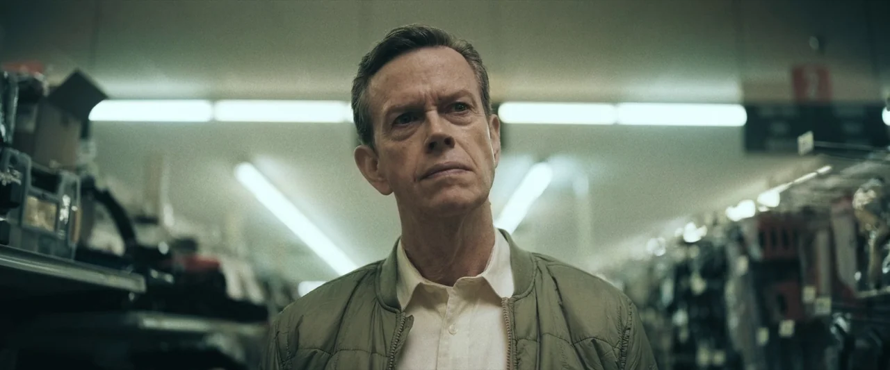 LaRoy, Texas (2023) movie still: medium — A man stands in the aisle of a retail store with blurred merchandise shelves. The man stares forward with…