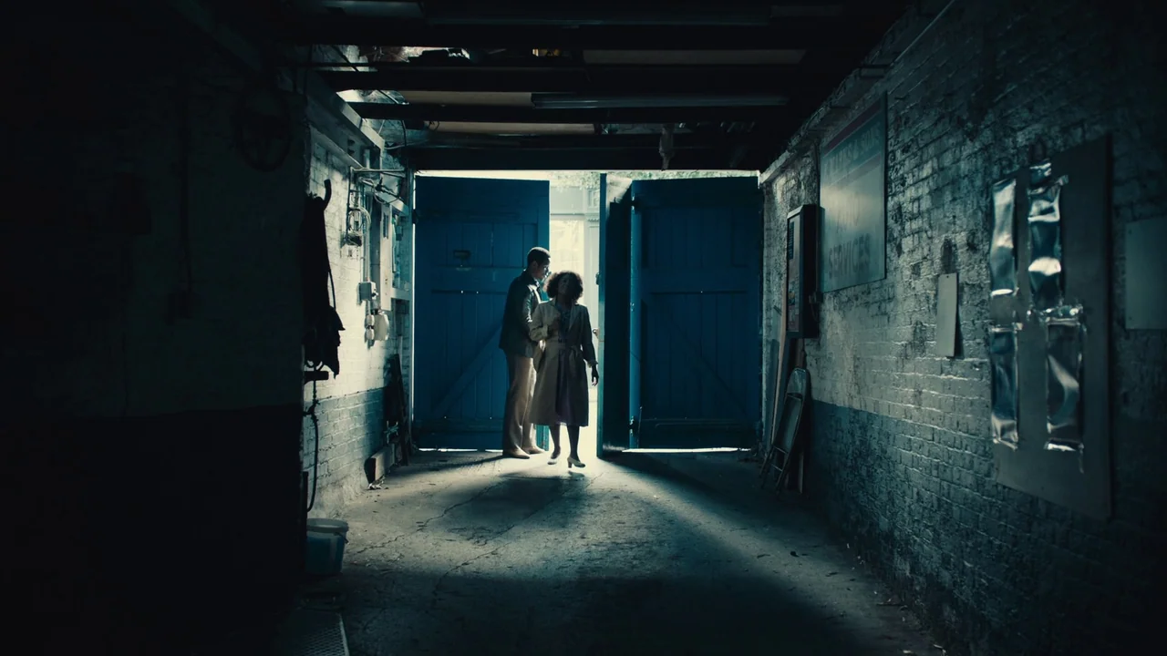 Lover’s Rock (2020) movie still: fullbody — A dimly lit industrial garage features whitewashed brick walls and heavy blue double doors. A man and a…