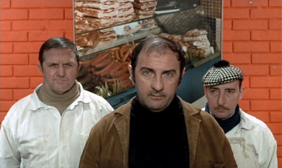 Tout Va Bien (1972) movie still: medium — A meat display case with sausages sits in front of a bright orange brick wall. Three men in professional…
