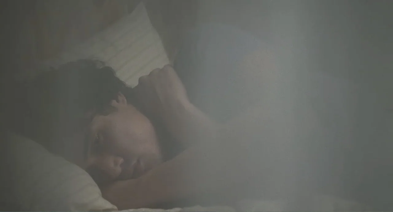 May December (2023) movie still: closeup — A dark bedroom interior shows a person resting on a bed under soft light. A sheer curtain partially…