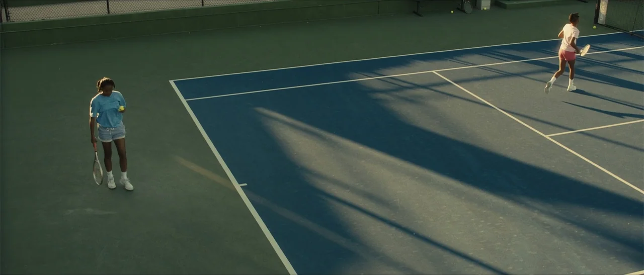 King Richard (2021) movie still: wide — Two girls stand on opposite sides of a blue hard-surface tennis court. One girl prepares to serve while the…