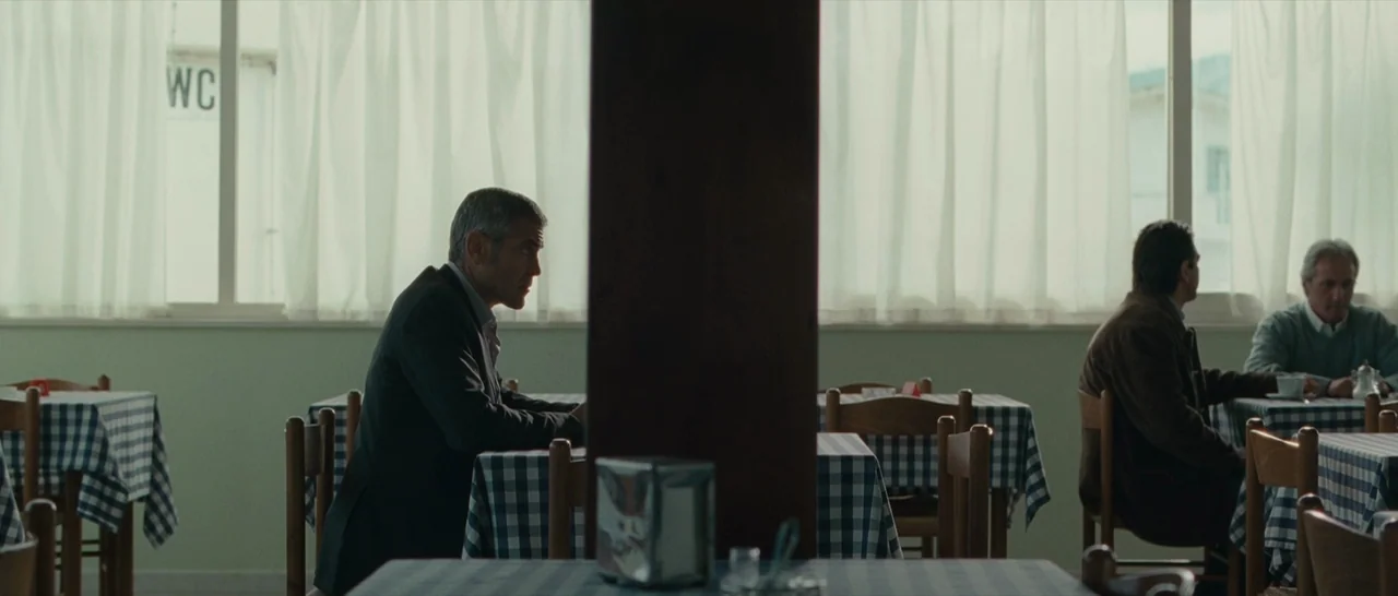 The American (2010) movie still: wide — A quiet cafe interior features blue checkered tablecloths and a large window with white curtains. A man sits…