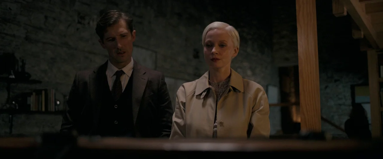 Oddity (2024) movie still: medium — A dimly lit room features a rugged stone wall background and wooden support beam. A man and woman stand close…