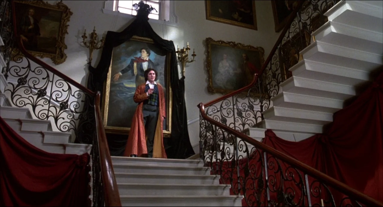 Gothic (1986) movie still: fullbody — A grand interior staircase features ornate iron railings and floor-to-ceiling red fabric drapes. A man stands…