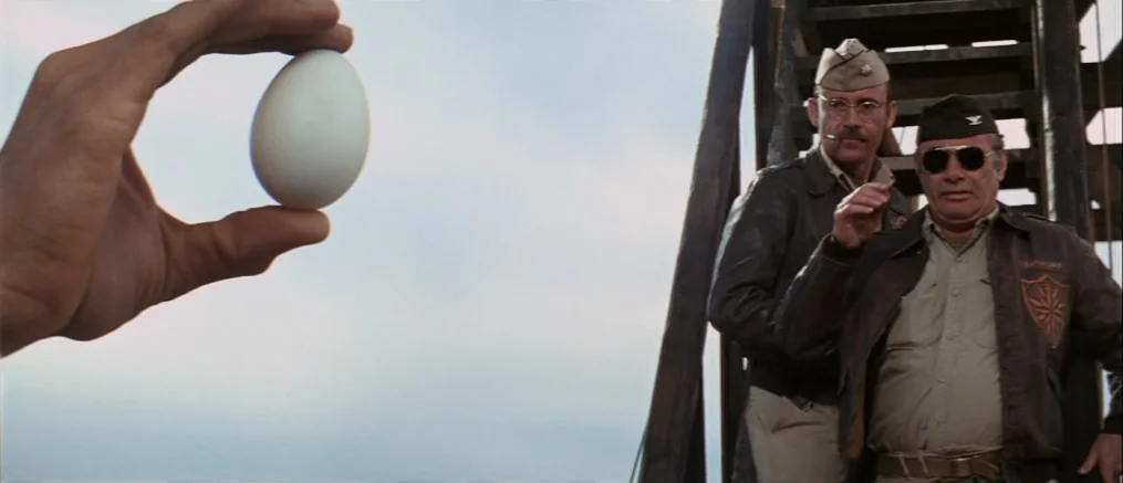 Catch 22 (1970) movie still: medium — Two military officers stand on a tall wooden training tower structure. A close-up foreground hand holds a…