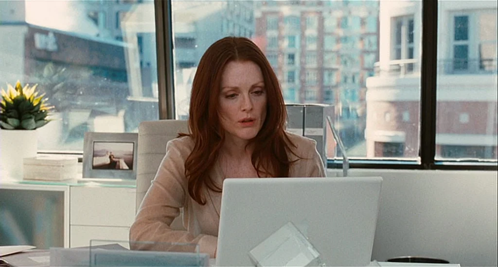 Chloe (2009) movie still: medium — A woman sits at a desk in a bright office with city views behind her. She looks intently at a laptop screen…
