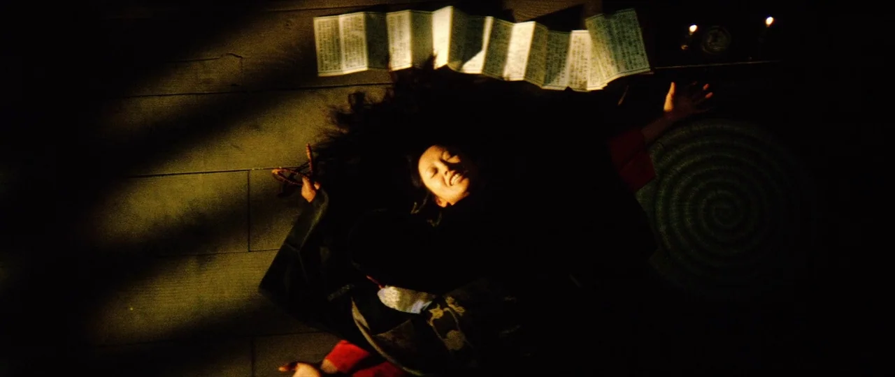Lady Snowblood (1973) movie still: medium — A dark, wood-floored room features a woman lying on the ground under intense light. An accordion-folded…
