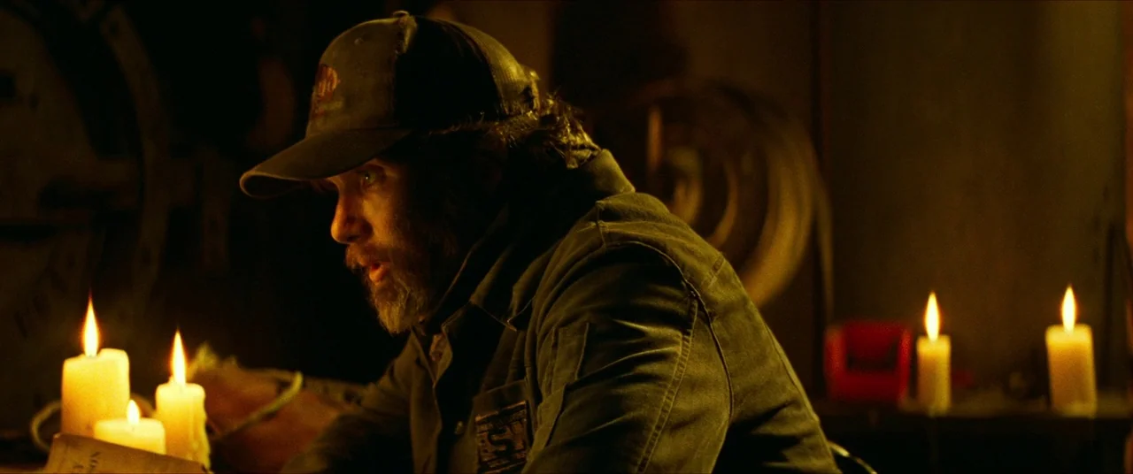 A Quiet Place Part II (2020) movie still: medium — A dimly lit room is filled with shadows and the warm glow of candles. A man wearing a work jacket…