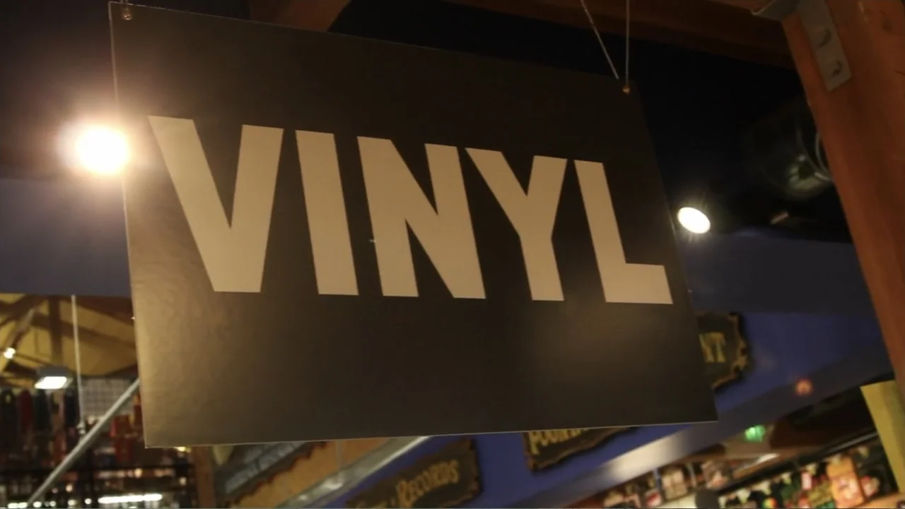 Hot Gimmick: Girl Meets Boy (2019) movie still: closeup — A record store interior features a prominent rectangular sign labeled VINYL hanging from…