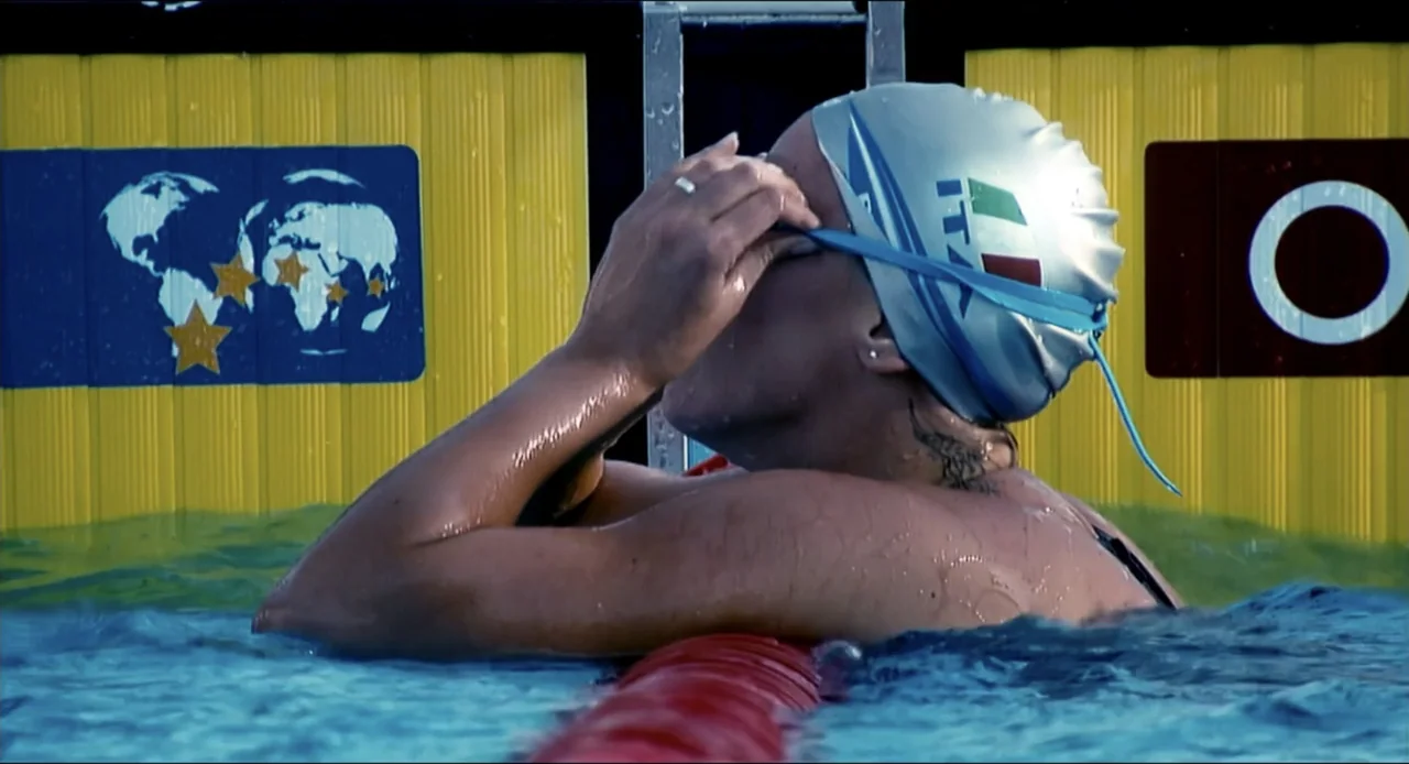 Federica Pellegrini - Underwater (2022) movie still: medium — A competitive swimmer rests at the edge of a bright blue outdoor pool. The athlete…