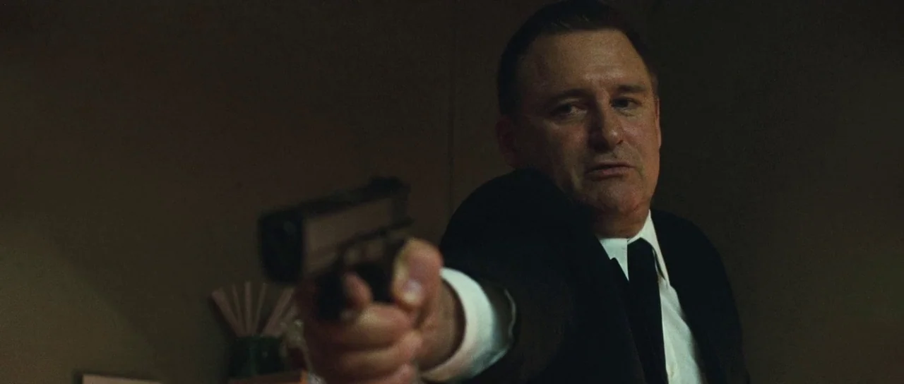 Surveillance (2008) movie still: medium — A dimly lit room with muted earth tones and deep shadows. A middle-aged man in a suit extends a pistol…