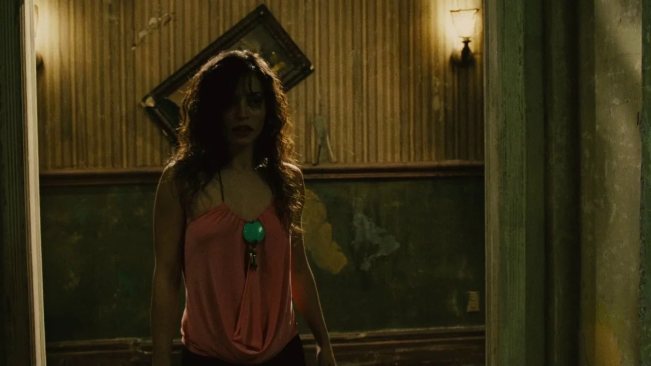 Saw II movie still: medium — A decrepit, dimly lit room features peeling wallpaper and a crooked picture frame. A woman wearing a pink top stands in…
