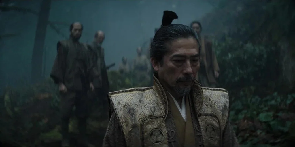 Shogun (2024) movie still: medium — A group of men moves through a dark, foggy forest with dense undergrowth. The foreground subject wears a…
