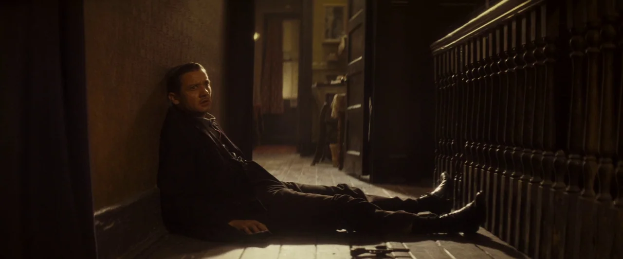 The Immigrant (2013) movie still: fullbody — A dimly lit narrow hallway with a wooden staircase balustrade running along one side. A man sits…