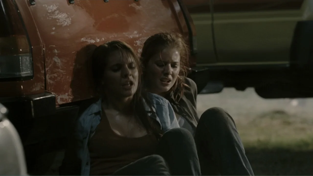 Tomorrow, When the War Began (2010) movie still: medium — A gravel surface outside an orange vehicle at night with two women sitting nearby. Two…