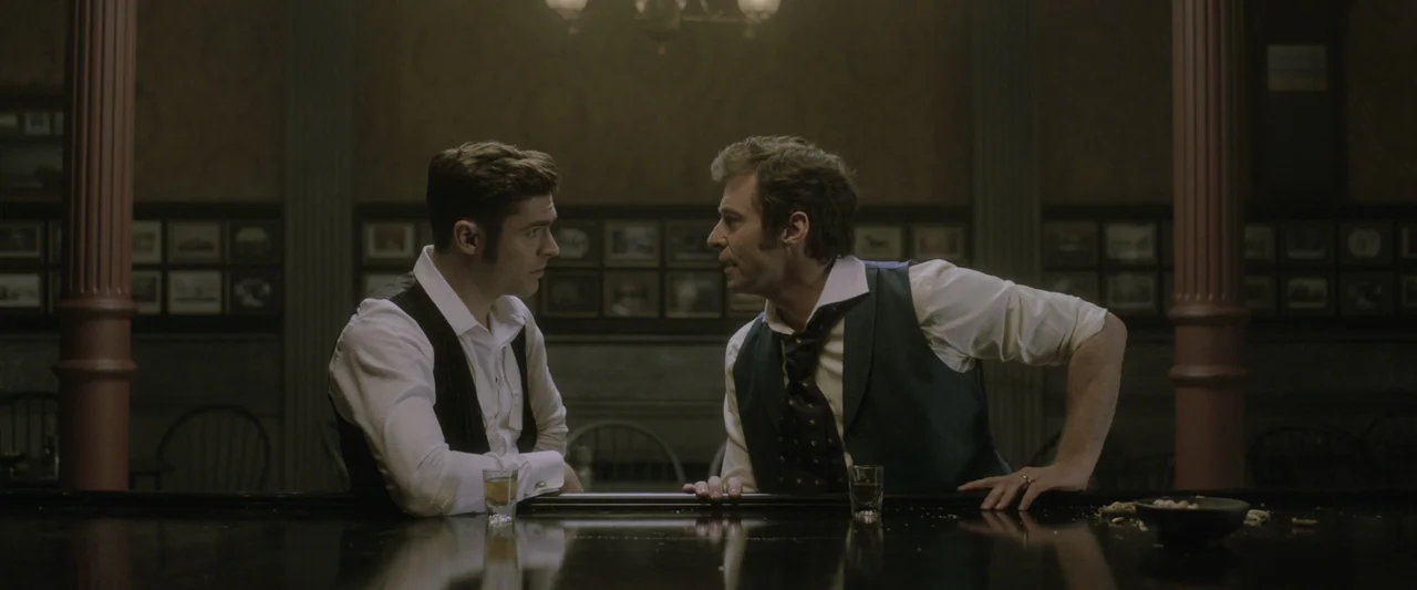 The Greatest Showman (2017) movie still: medium — A dimly lit 19th-century pub with dark wood paneling and decorative wall frames. Two men in period…