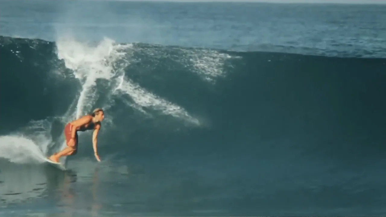 El sueño (2020) movie still: wide — A dark blue breaking wave curves across the surface of the open ocean. A lone surfer maneuvers a surfboard along…