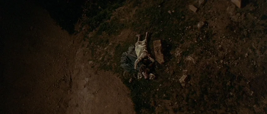 Flesh+Blood (1985) movie still: wide — An uneven dirt slope at night with patches of sparse grass and loose rocks. A woman lies face down on the…