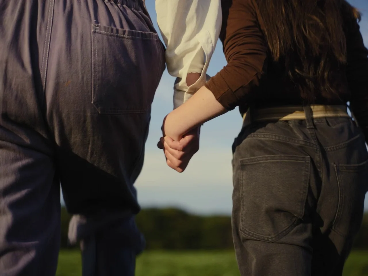 The Quiet Girl (2022) movie still: medium — Two individuals stand in a grassy field with a clear blue sky above. They are holding hands while viewed…