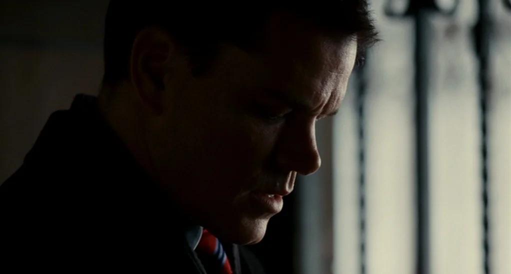 The Adjustment Bureau (2013) movie still: closeup — The man stands indoors against a blurred vertical window or gate. He looks downward with his…