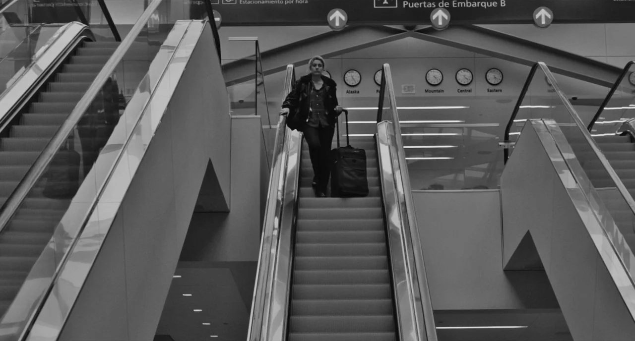 Frances Ha (2012) movie still: fullbody — An airport terminal with multiple escalators and wall clocks showing different time zones. A woman pulls a…