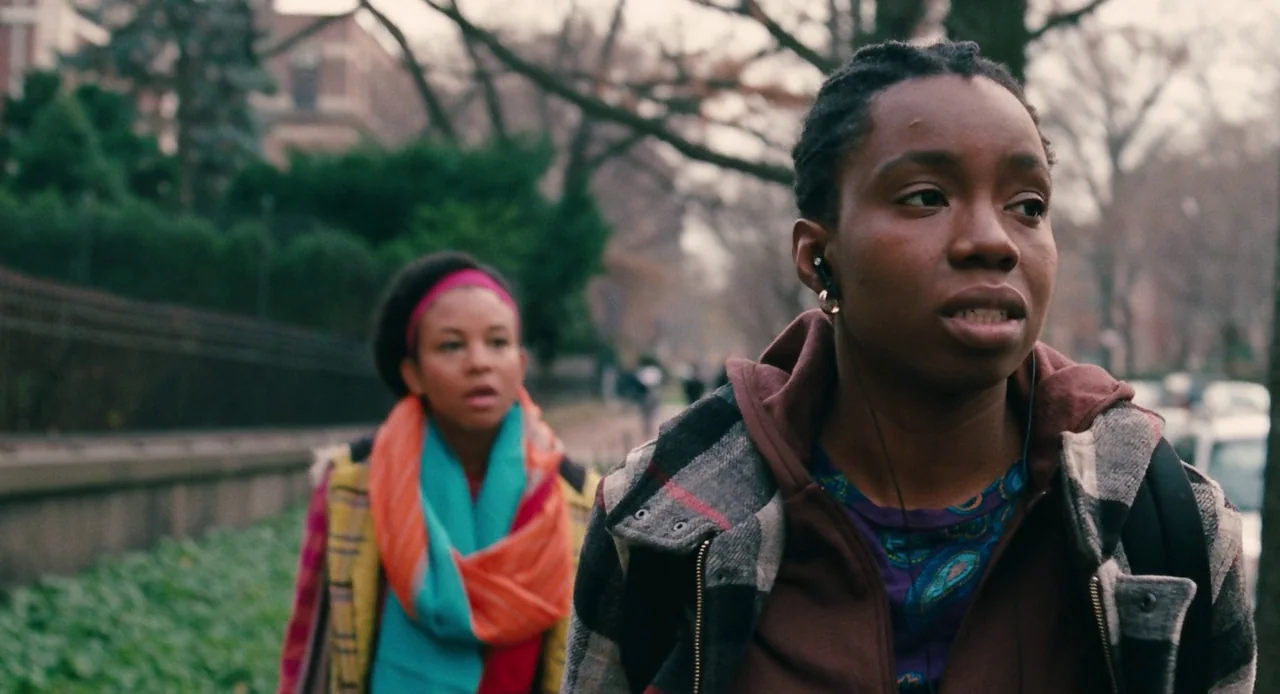 Pariah (2011) movie still: medium — A sidewalk path bordering a brick building and bare trees during autumn or winter. Two young women walk…