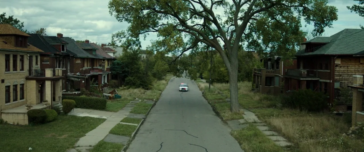 Don’t Breathe (2016) movie still: wide — A residential street is lined with weathered, aging brick houses and overgrown lawns. A single police…
