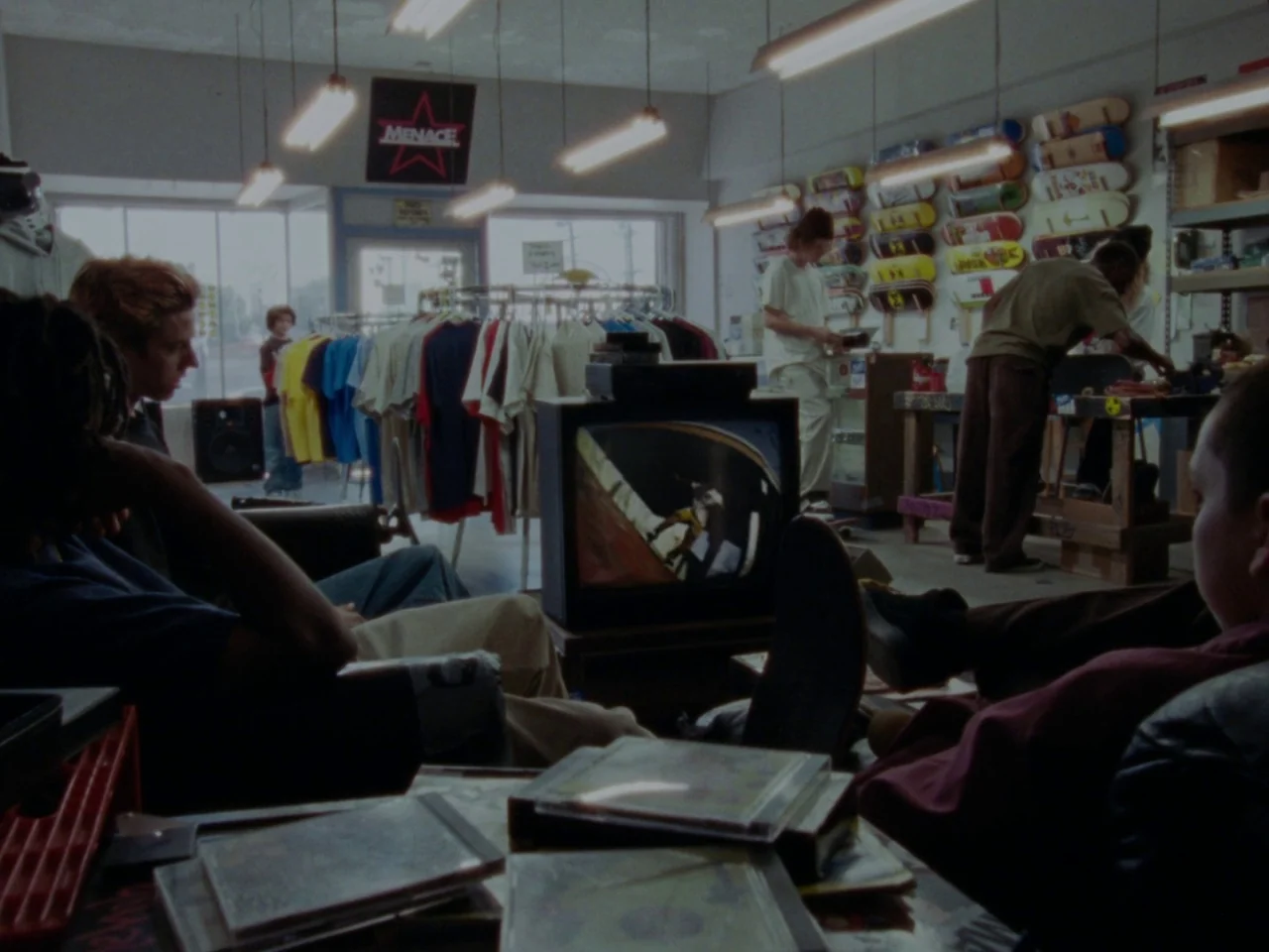 Mid90s (2018) movie still: wide — A cluttered 1990s retail space filled with skate decks on racks and clothing. Several teenagers sit in a casual…