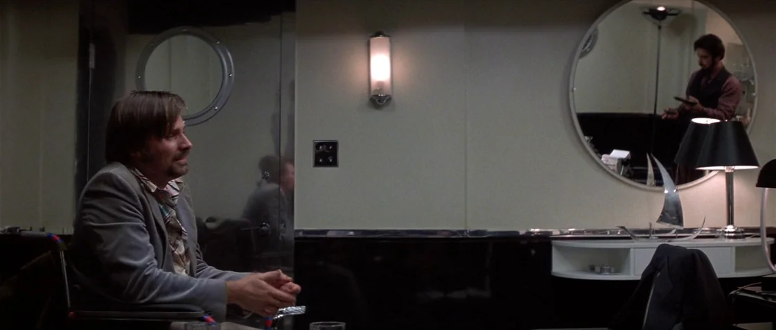 Carlito’s Way (1993) movie still: medium — An interior space features reflective dark walls and a circular wall mirror reflecting a man. A man sits…