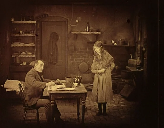 Broken Blossoms (1919) movie still: fullbody — A rustic room with brick walls and shelves containing various household items. A man sits at a wooden…
