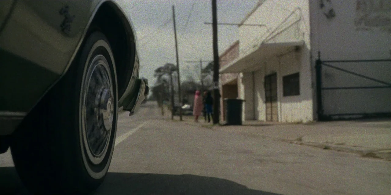 They Cloned Tyrone (2023) movie still: wide — An asphalt road runs alongside a weathered white building with a small awning. Two indistinct figures…