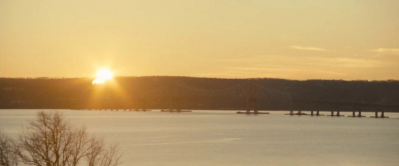 The Skeleton Twins (2014) movie still: establishing — A wide view shows a large bridge spanning across a calm body of water. The sun sets behind…