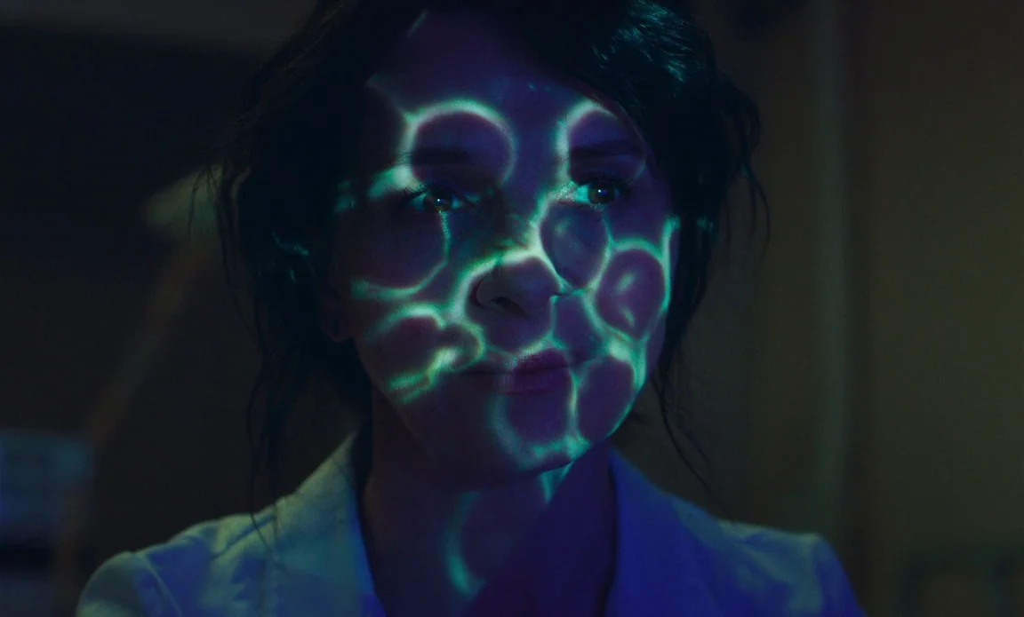 High Life (2018) movie still: closeup — A dimly lit room features a woman with light patterns projected across her face. She stares forward while…