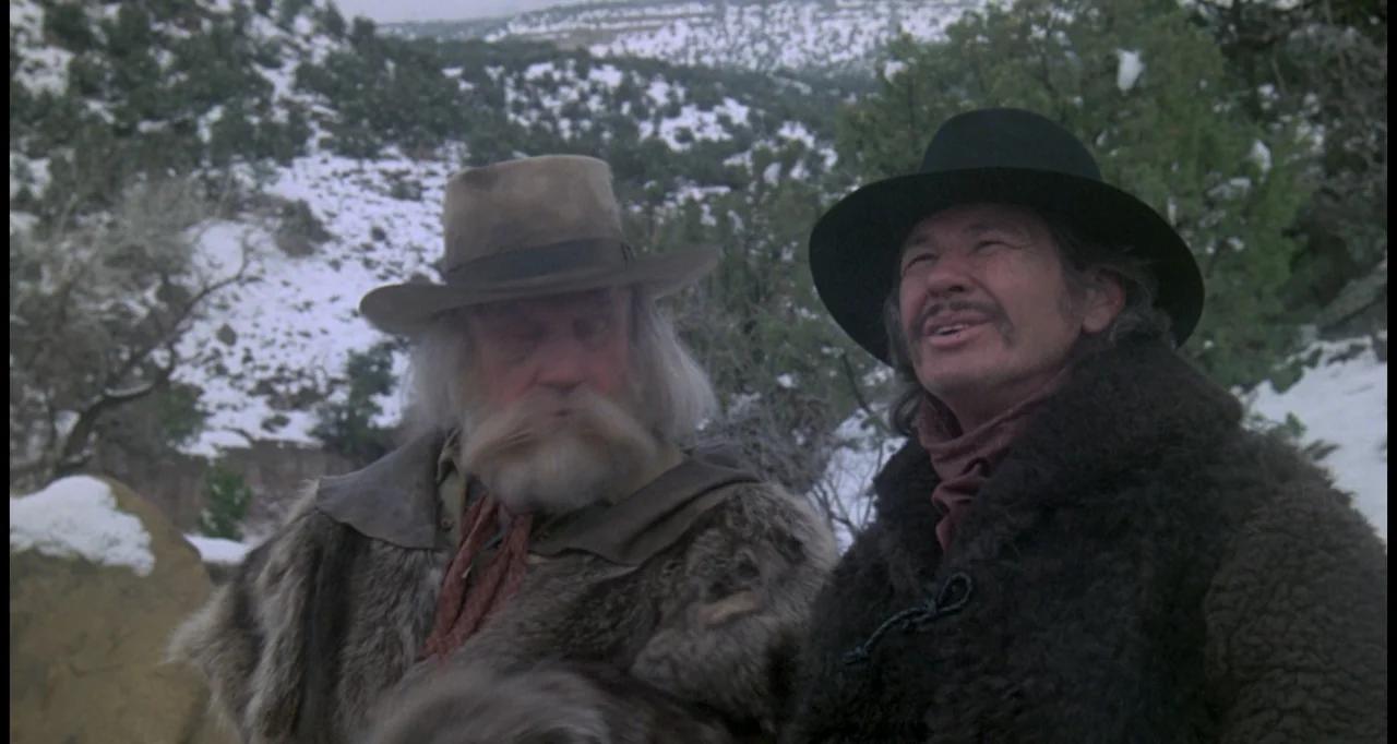 The White Buffalo (1977) movie still: medium — A snowy, brush-covered mountain slope provides the background for two men. Two men dressed in heavy…
