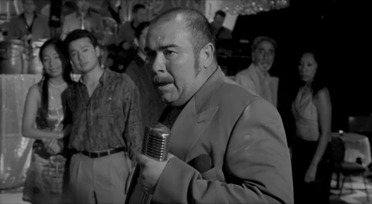 Sex, Love And Other Perversions II (2006) movie still: medium — A dimly lit nightclub features a band and a small audience in the background. A man…