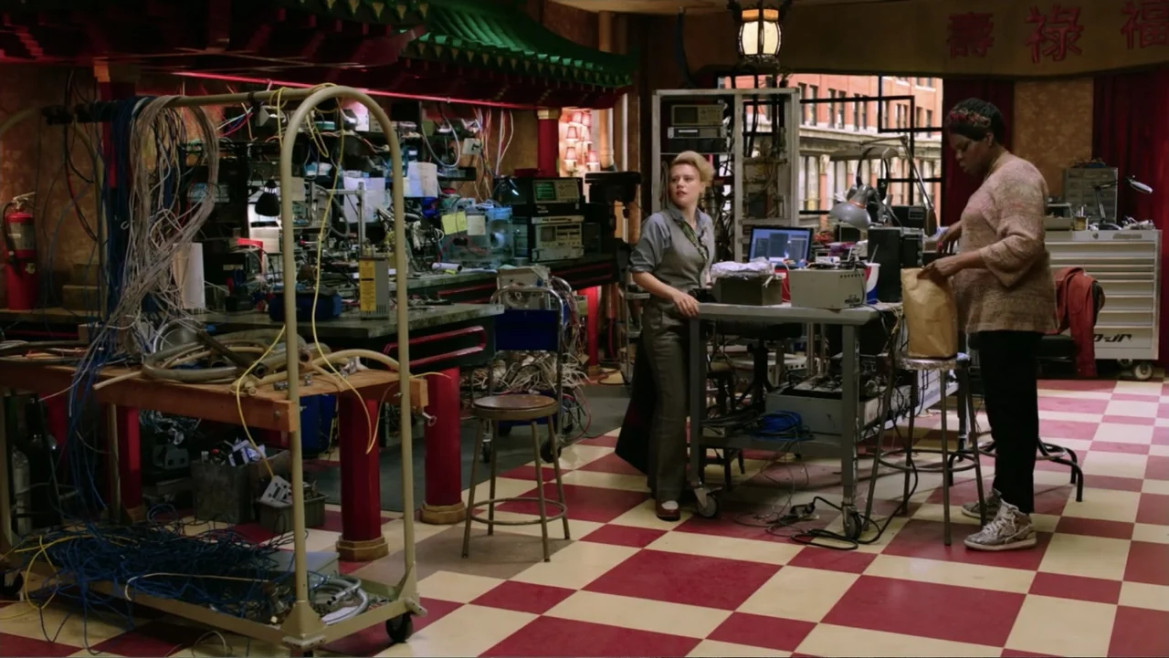 Ghostbusters (2016) movie still: fullbody — A cluttered workshop features a red and white checkered tile floor and electronic equipment. Two women…
