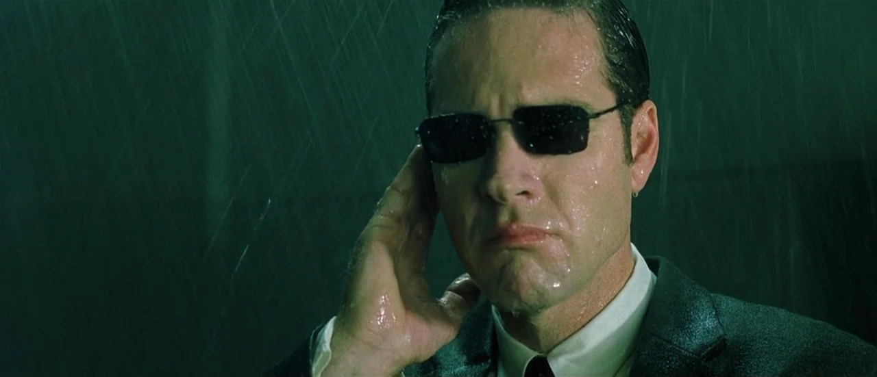 The Matrix (1999) movie still: closeup — A man stands in the pouring rain during a dark night in the city. He holds his hand up toward his ear while…