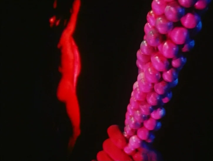 Pink Narcissus (1971) movie still: closeup — A dark environment displays a cluster of pink spheres in the immediate foreground. In the blurry…