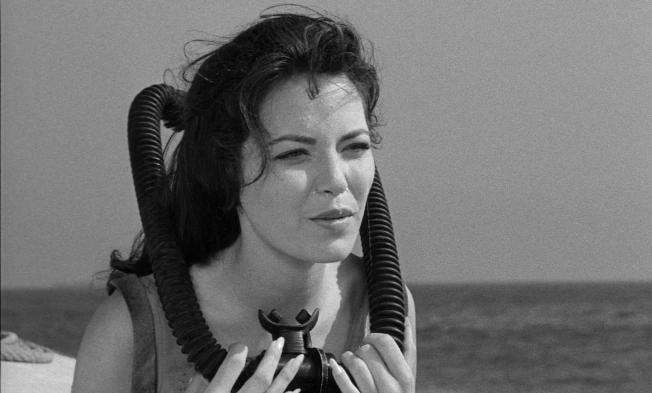 Night Tide (1961) movie still: medium — A woman stands on a boat deck with the vast ocean behind her. She holds a black scuba regulator hose around…
