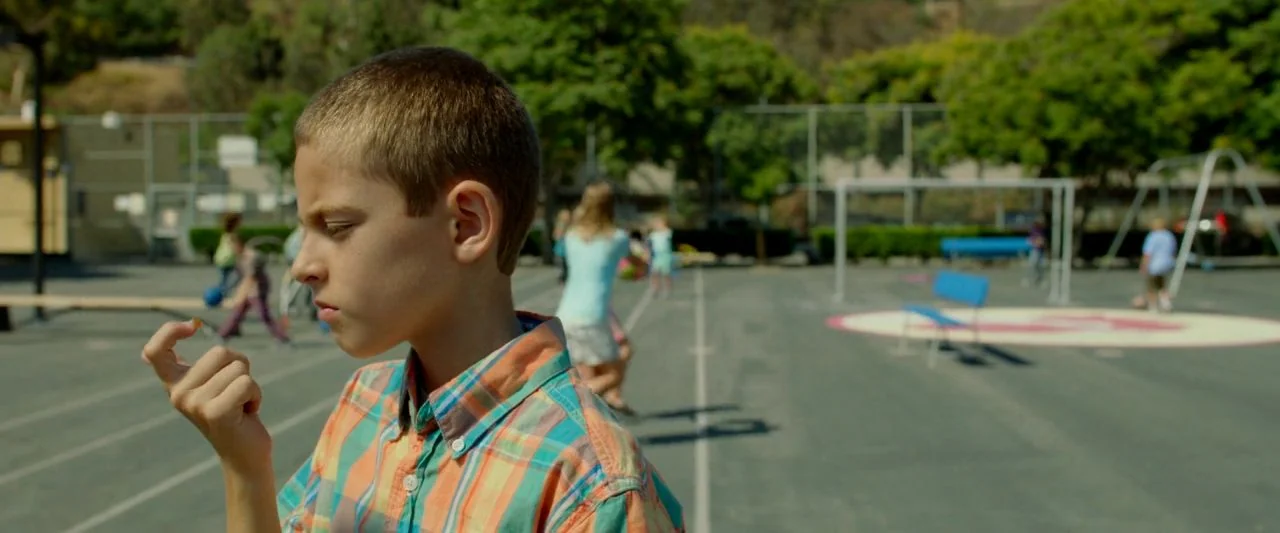 Cooties (2014) movie still: medium — A paved playground at a school with trees and a metal fence in distance. A young boy looks at a small object he…