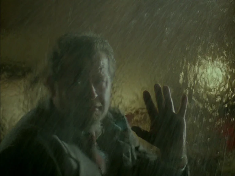 Helas Pour Moi (1993) movie still: closeup — A man stands behind a heavily rain-streaked glass surface at night. He presses his open palm against…