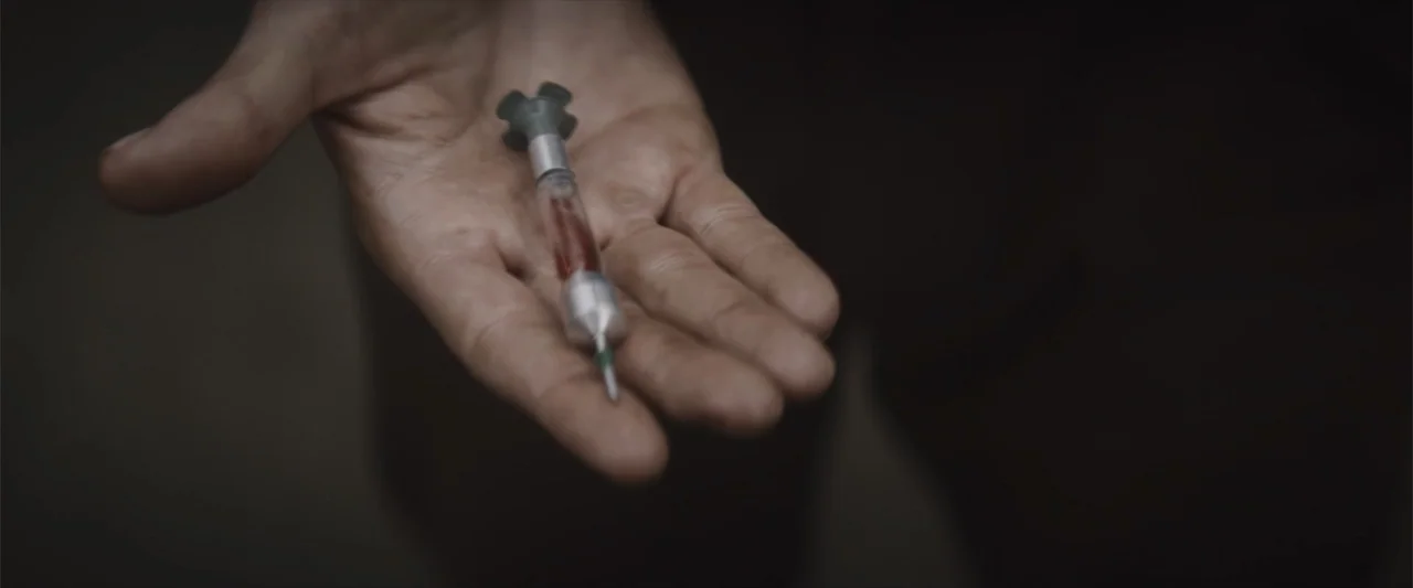 Treblinka's Last Witness (2012) movie still: closeup — An open palm holds a metallic medical syringe containing a red liquid. The background is a…