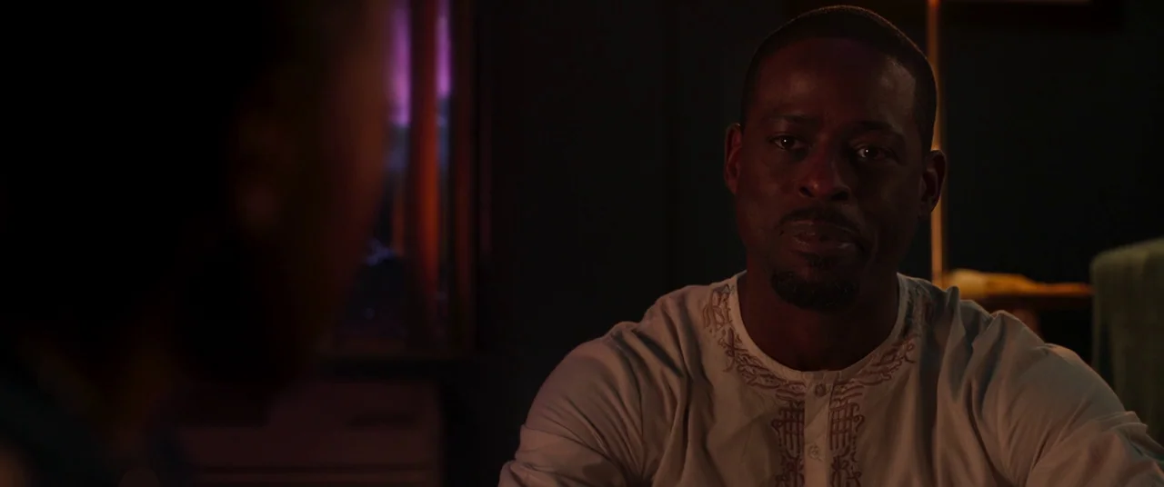 Black Panther (2018) movie still: medium — A dimly lit interior room at night contains a man sitting in the foreground. He is listening to someone…