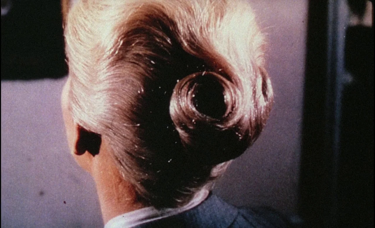 Sans Soleil (1983) movie still: closeup — A close view of a woman with her blonde hair pulled into a bun. The subject turns her head while facing…