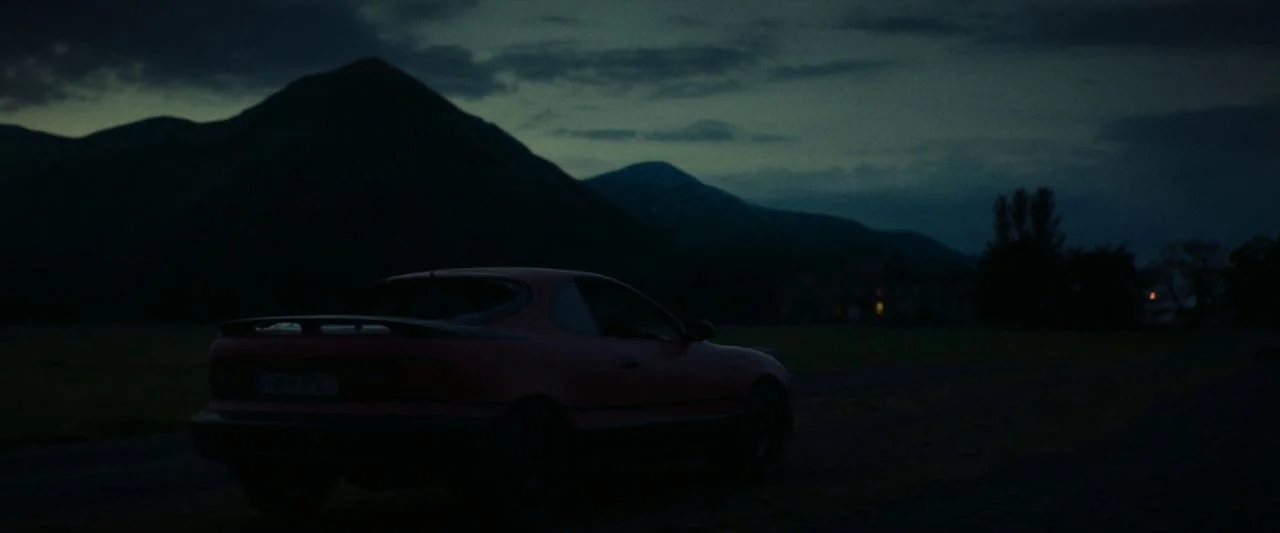 Calm With Horses (2019) movie still: wide — A red sports car sits parked in a dark, open rural field at dusk. Large silhouetted mountains rise…