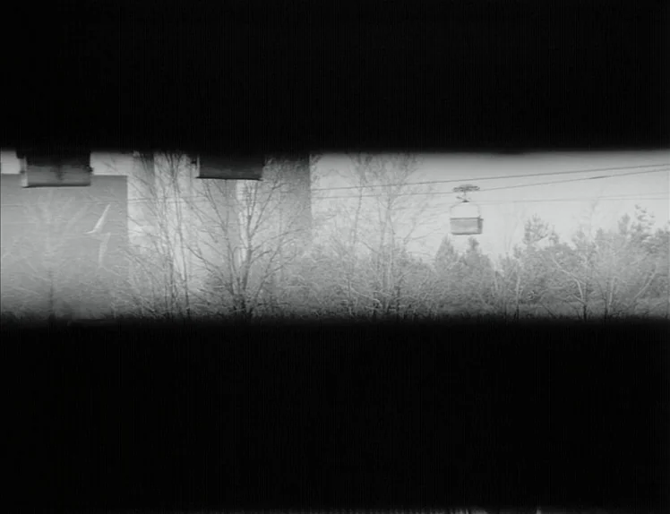 Damnation (1988) movie still: establishing — A view through a narrow horizontal slit shows a landscape with bare trees. A lone chairlift car hangs…
