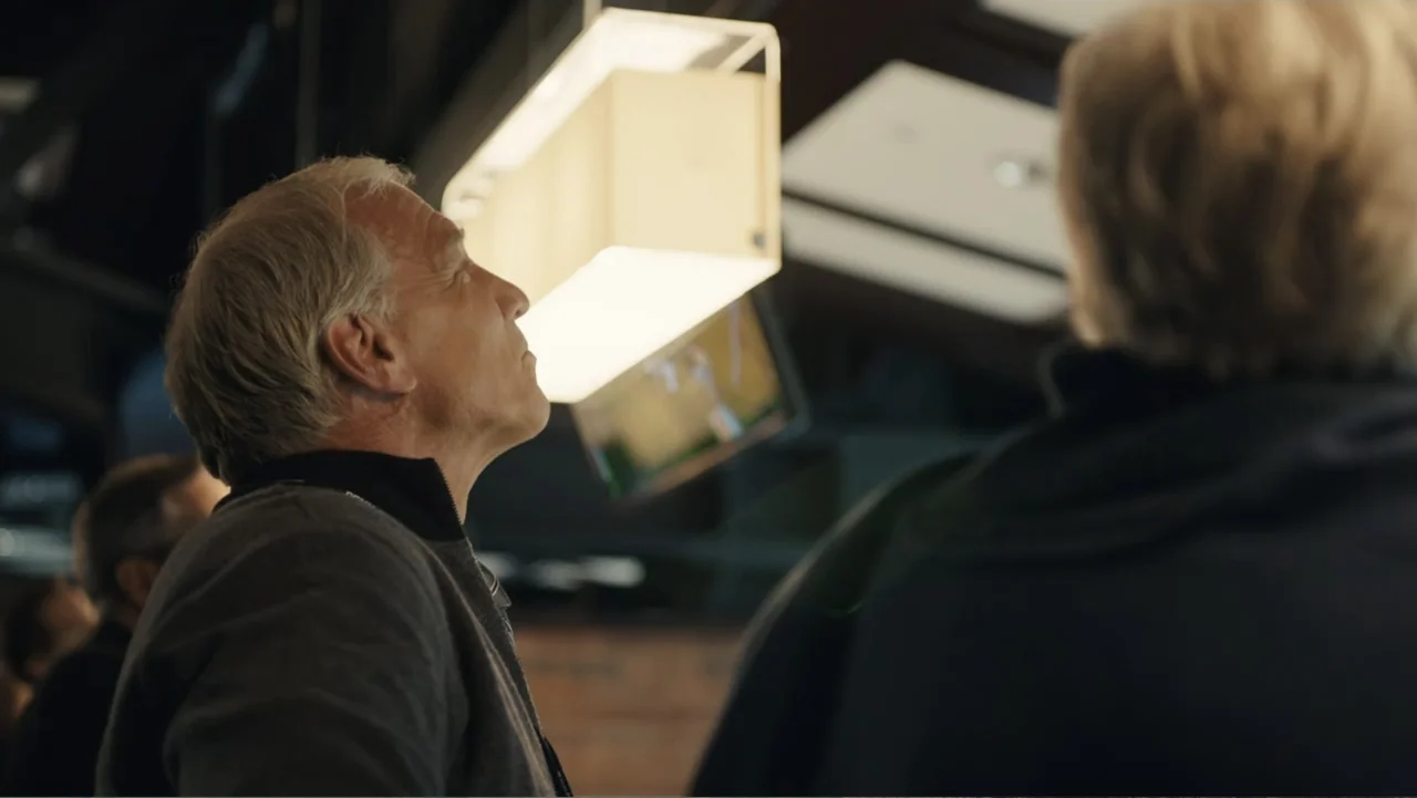 Paterno (2018) movie still: medium — A dimly lit indoor space features a prominent rectangular hanging light fixture. Two individuals stand looking…