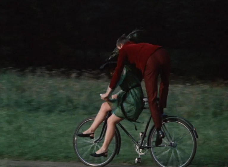 Fruit of Paradise (1970) movie still: fullbody — A grassy meadow serves as the backdrop for a pair riding a bicycle. The woman steers while the man…