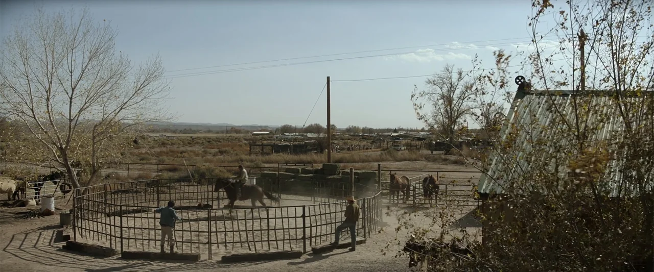 Cry Macho (2021) movie still: wide — An outdoor dusty riding arena sits in a rural desert landscape with scattered vegetation. One individual on a…