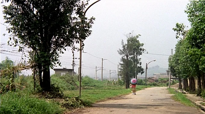 A Summer at Grandpa’s (1984) movie still: wide — A quiet, paved road stretches between dense greenery and overhead power lines. A small child walks…